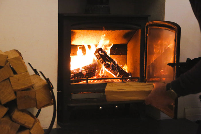 A log burner containing a roaring fire. The door is open and someone is putting a new log on the fire.